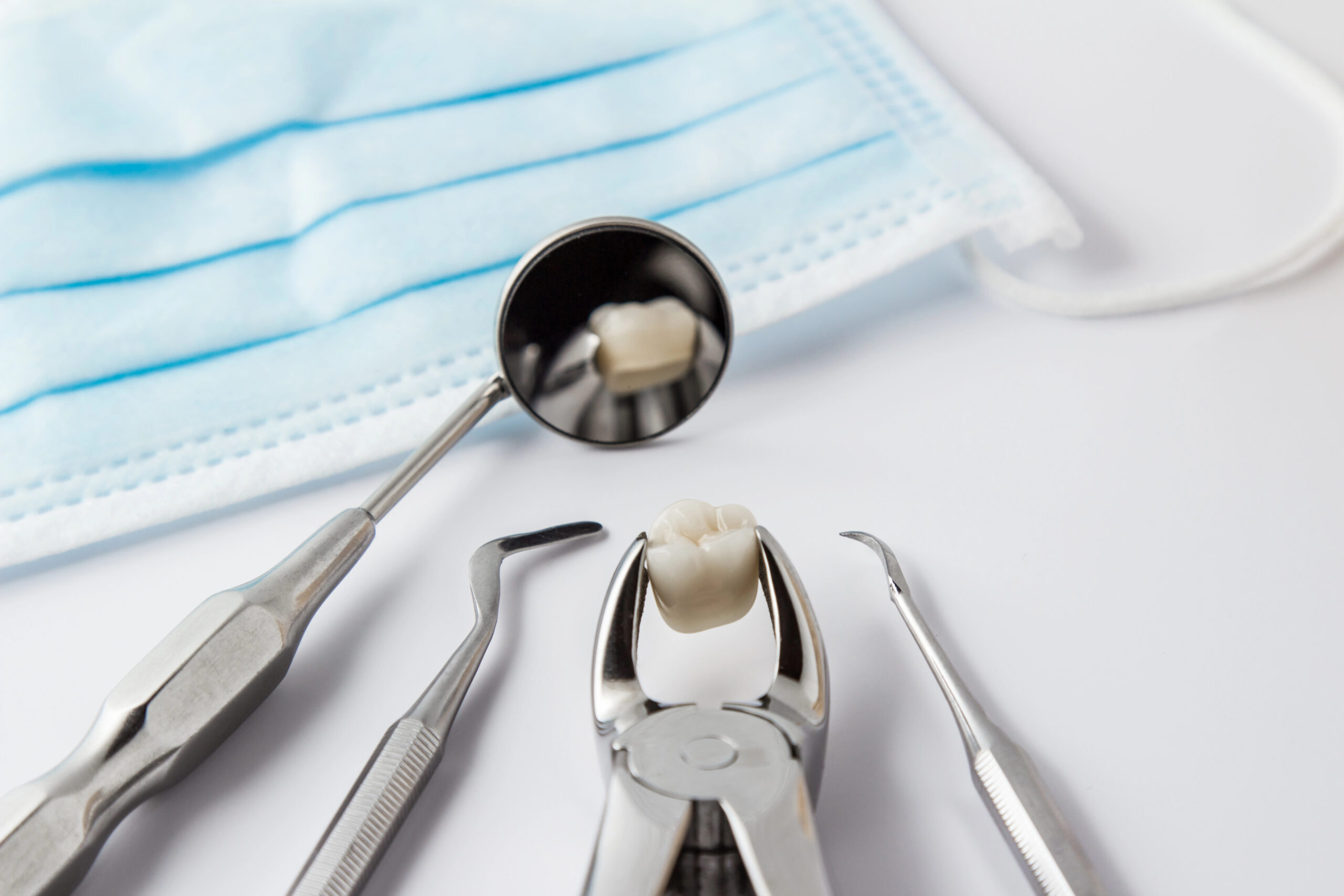 A set of dental tools, including forceps holding an extracted tooth, displayed alongside a dental mirror and mask, representing the process of wisdom teeth removal without sedation in a clinical setting.